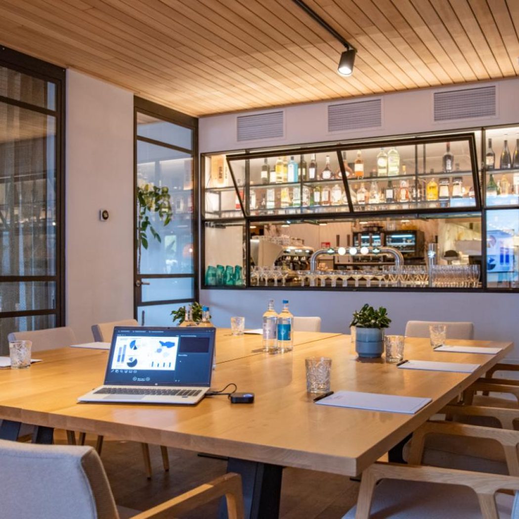 Stijlvolle vergaderruimte met houten tafel, laptop die een presentatie toont, en een bar achter glas.