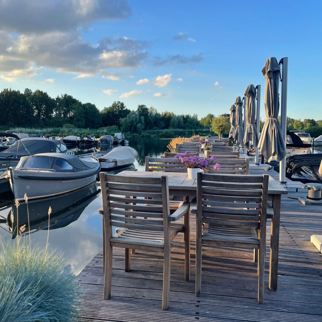 Zonnig houten terras met tafels, stoelen, opgevouwen parasols en aangemeerde boten bij een jachthaven.