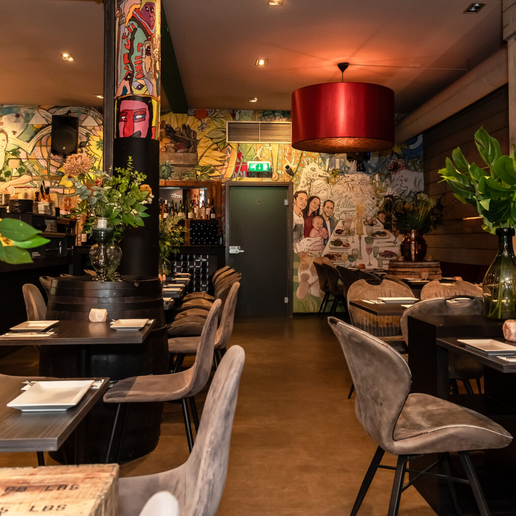Warm verlicht restaurantinterieur met kleurrijke muurschilderingen, donkere tafels, grijze stoelen en groene plantdecoraties.