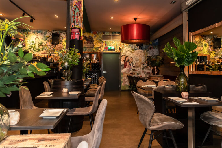 Warm verlicht restaurantinterieur met kleurrijke muurschilderingen, donkere tafels, grijze stoelen en groene plantdecoraties.