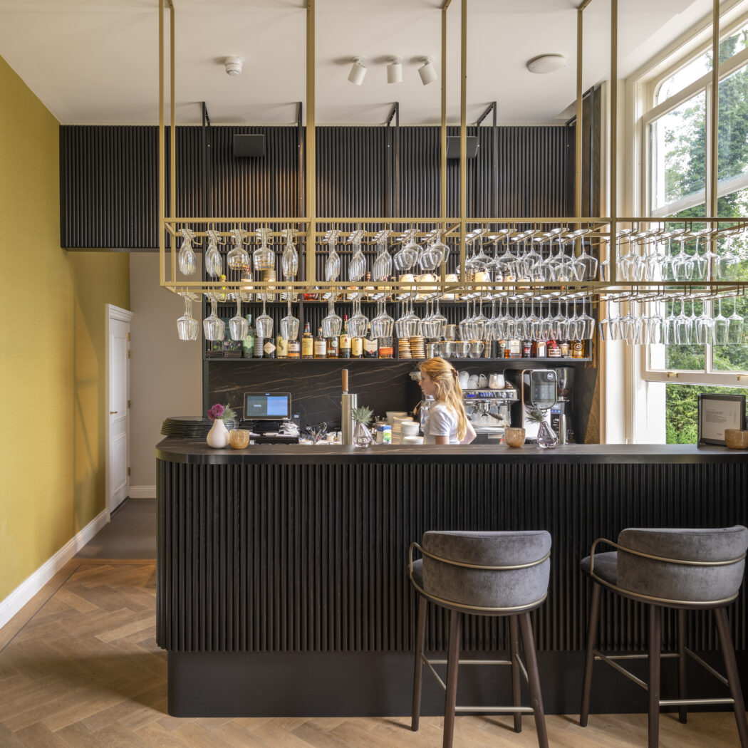 Elegante bar met donkere lamellen, gouden glasrek en barista, naast een mosterdgele muur en raam met groen uitzicht.