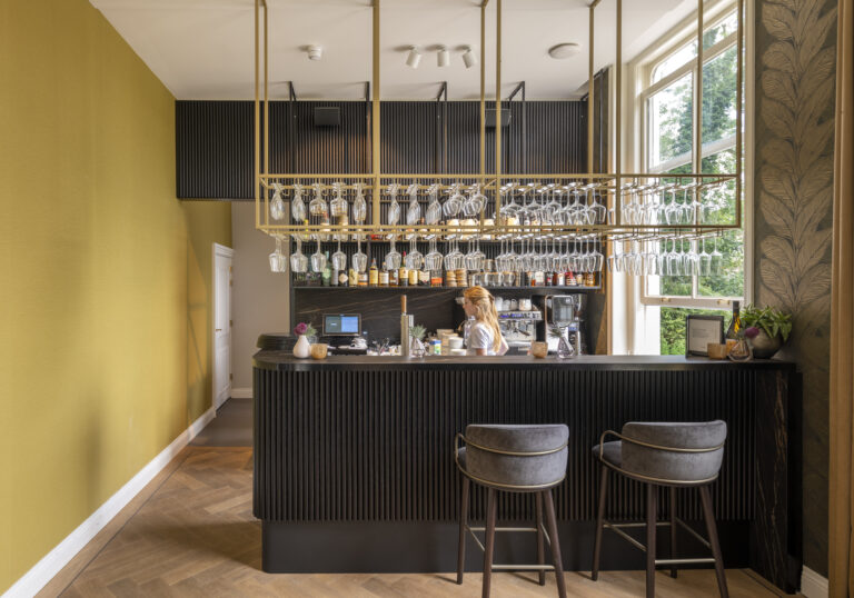 Elegante bar met donkere lamellen, gouden glasrek en barista, naast een mosterdgele muur en raam met groen uitzicht.