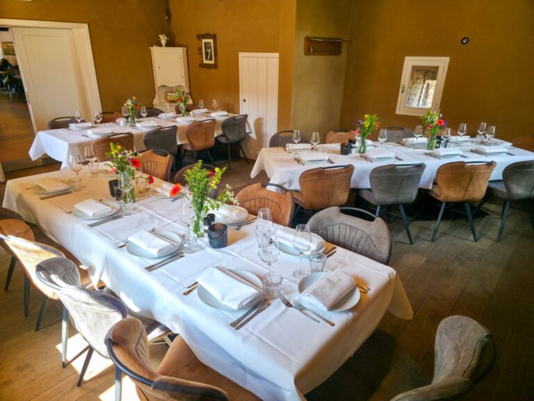 Gezellige restaurantzaal met lange gedekte tafels, witte kleden, bloemstukken en Sanora-servetten.