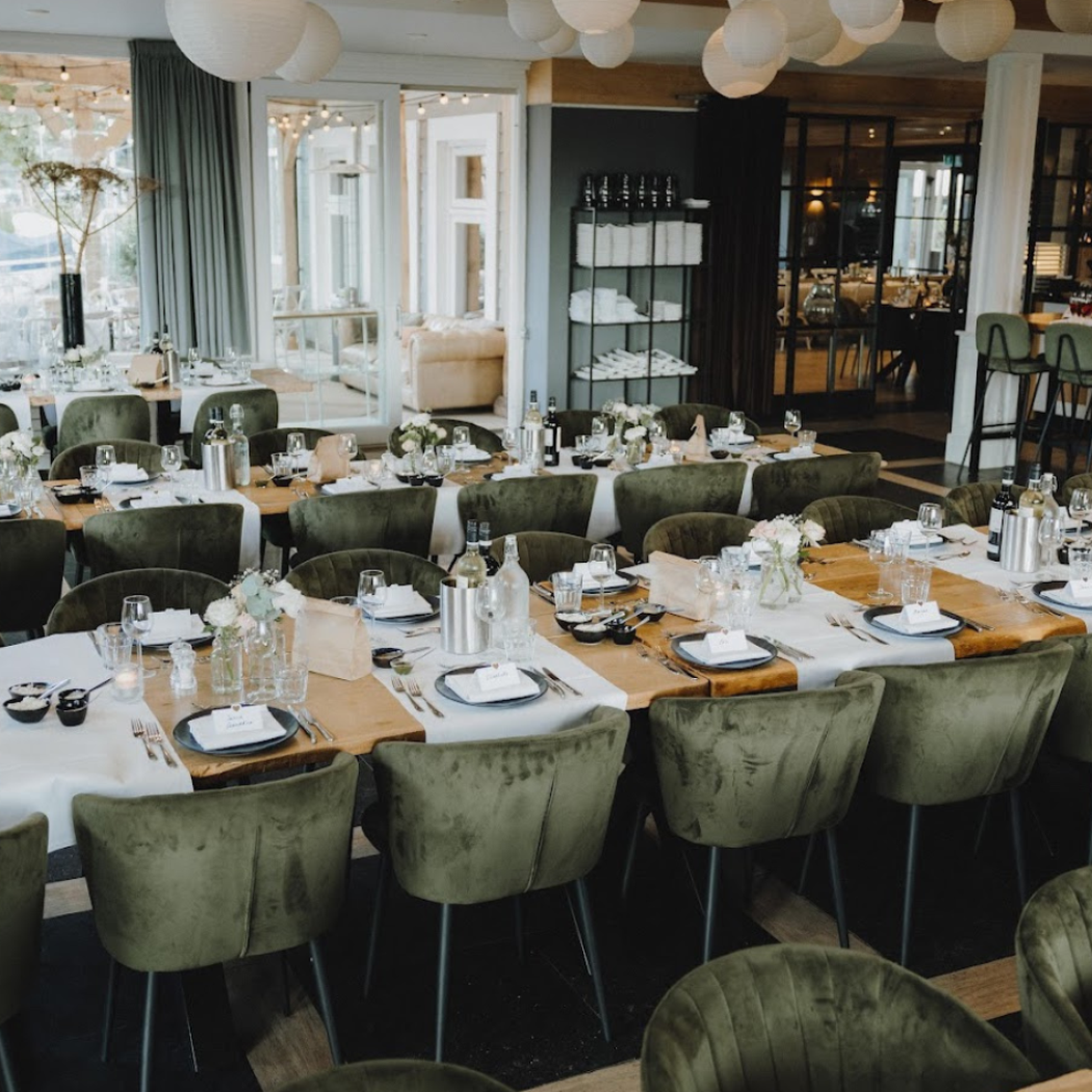 Sfeervolle evenementenlocatie met gedekte houten tafels, groene fluwelen stoelen en een waterrijk uitzicht.