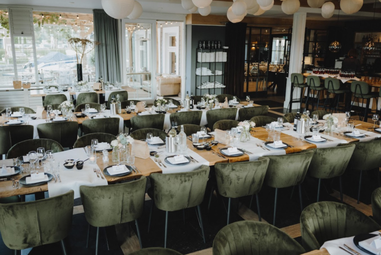 Sfeervolle evenementenlocatie met gedekte houten tafels, groene fluwelen stoelen en een waterrijk uitzicht.