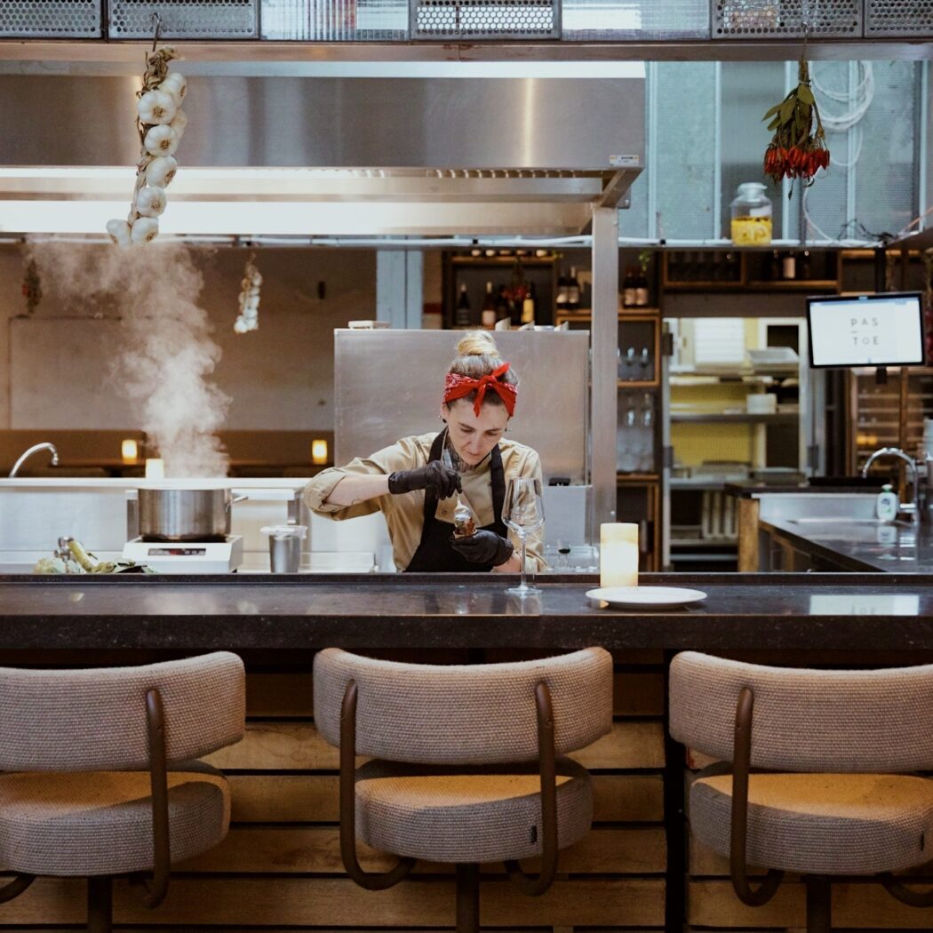 Chef met rode bandana en zwarte handschoenen bereidt een gerecht achter bar, stoom uit pan, met PAS TOE scherm.