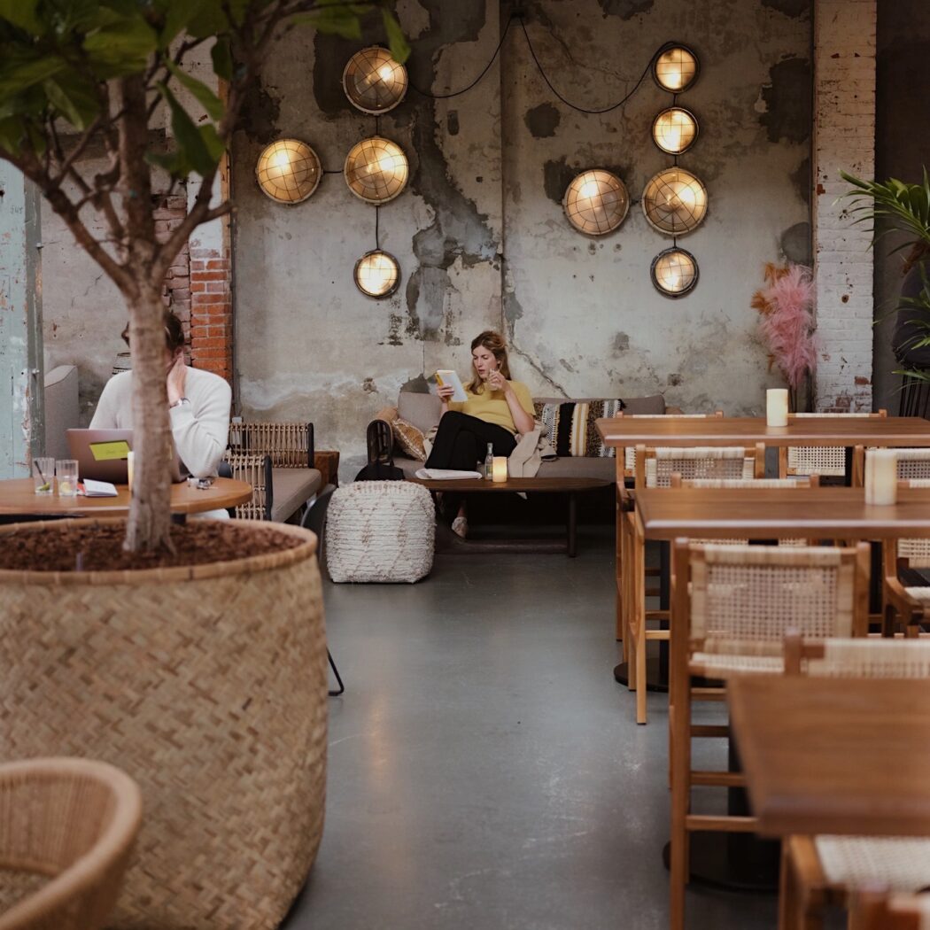 Vrouw leest een boek op een bank in een café met industriële wandlampen en een grote plant in een rieten pot.