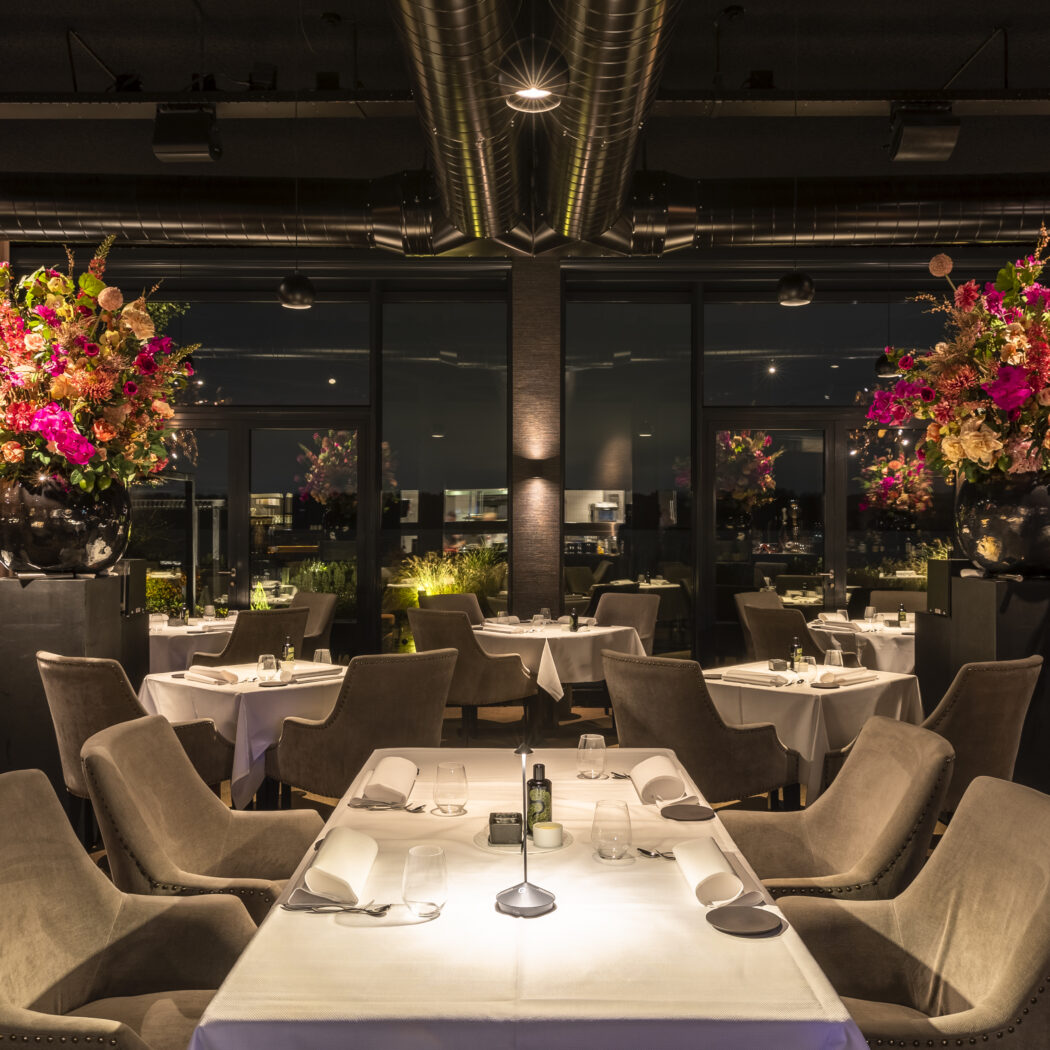 Elegant restaurantinterieur s avonds met gedekte tafels, bruine stoelen en twee grote, kleurrijke bloemstukken.