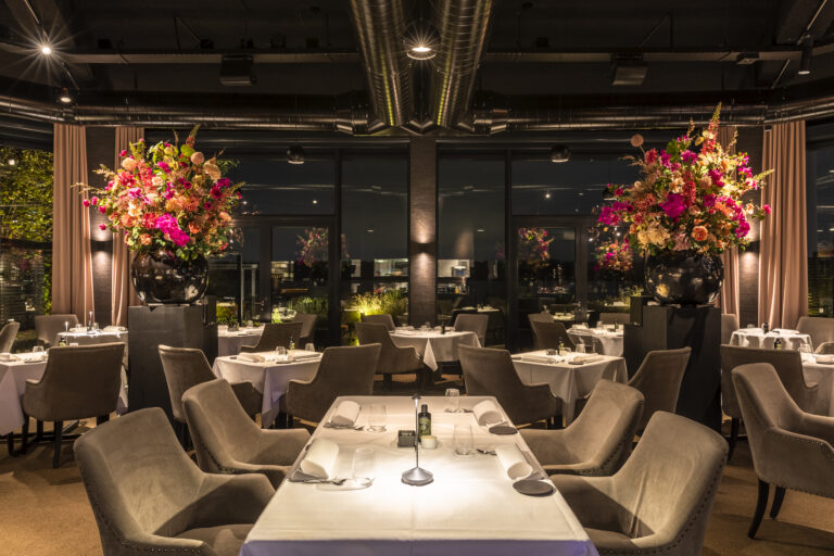 Elegant restaurantinterieur s avonds met gedekte tafels, bruine stoelen en twee grote, kleurrijke bloemstukken.