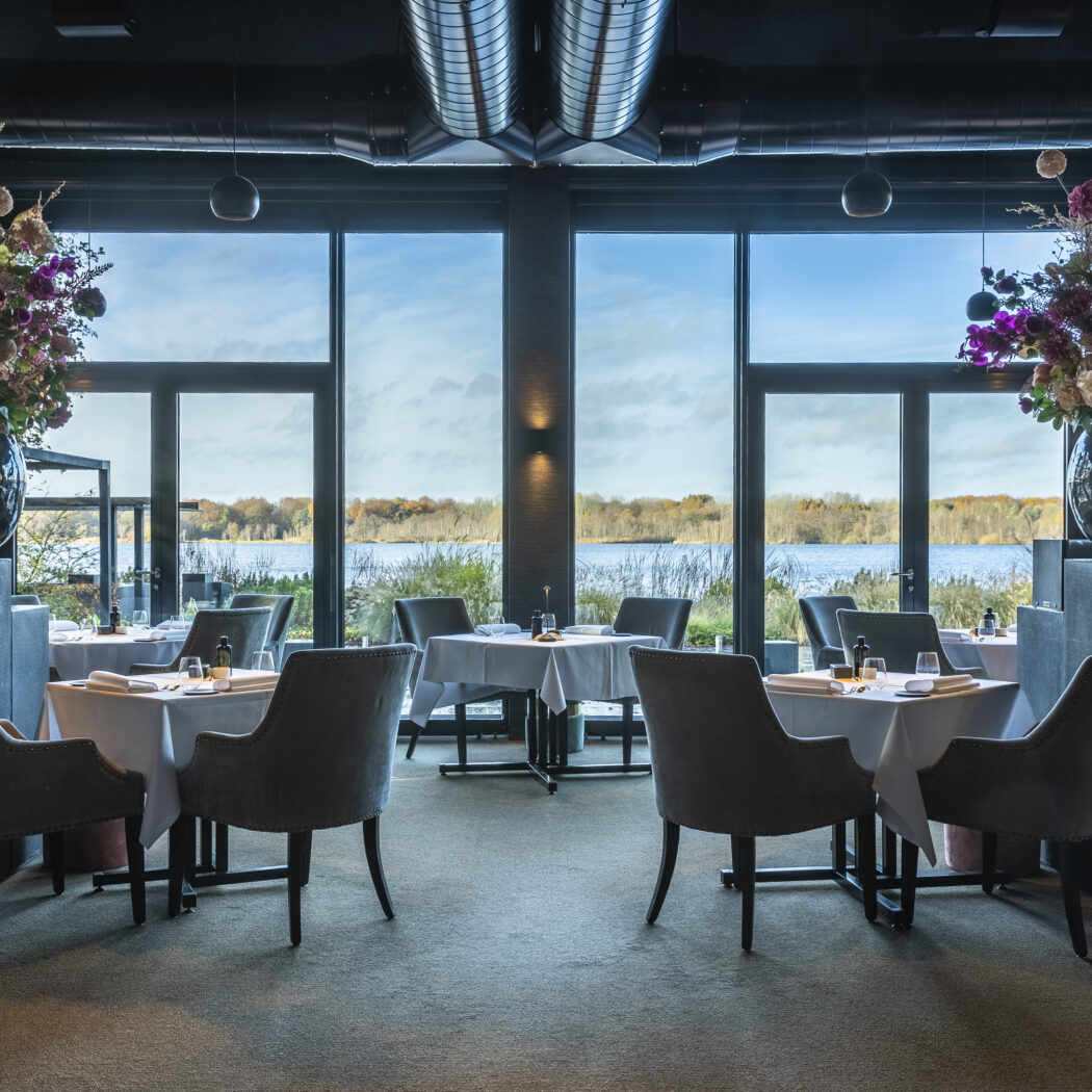 Elegant restaurantinterieur met gedekte tafels, grijze stoelen, grote bloemstukken en uitzicht op water.