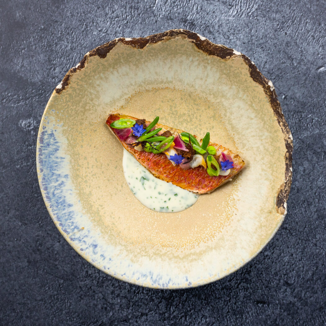 Rode visfilet met groene kruiden, blauwe bloemetjes en romige saus op een handgemaakt aardewerk bord.