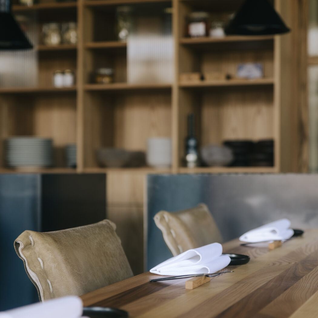 Houten eettafel in een restaurant, gedekt met witte servetten en donkere placemats, met beige stoelen en een kast vol items.