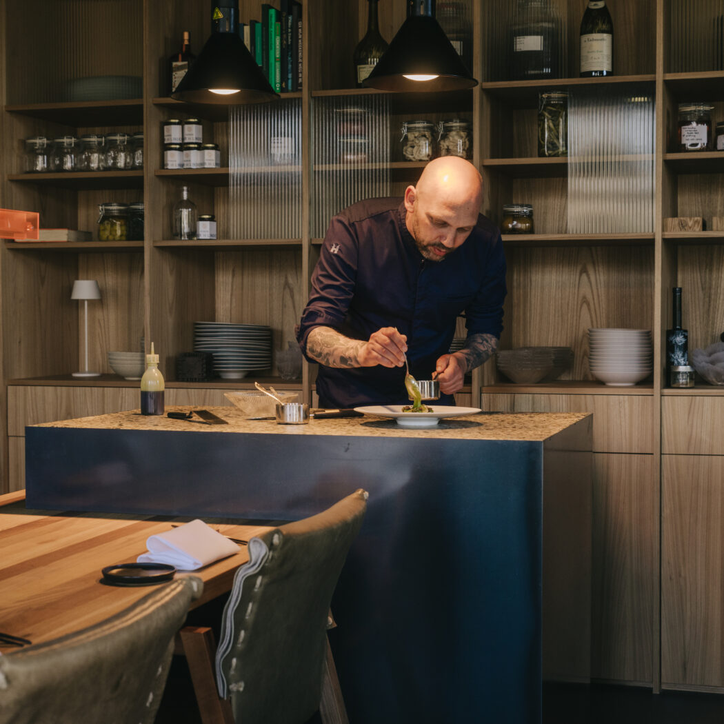 Chef met tatoeages maakt een gerecht op met groene garnituur in een stijlvolle keuken.