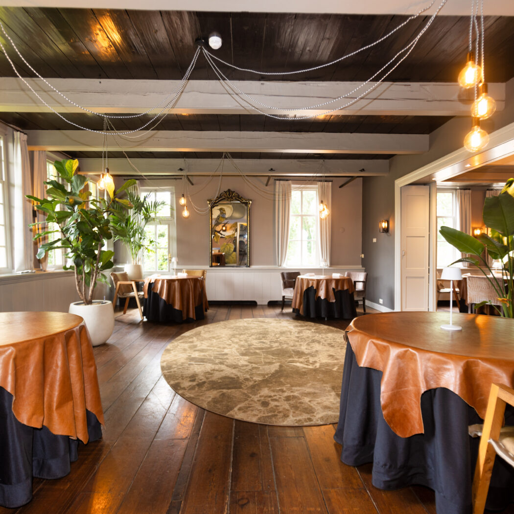 Ruime restaurantzaal met bruine ronde tafels, houten vloer, donkere balken, hanglampen en grote groene planten.