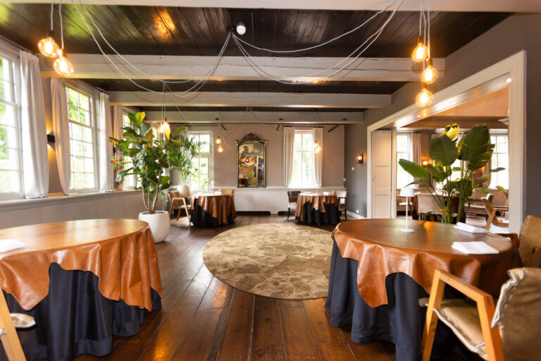 Ruime restaurantzaal met bruine ronde tafels, houten vloer, donkere balken, hanglampen en grote groene planten.