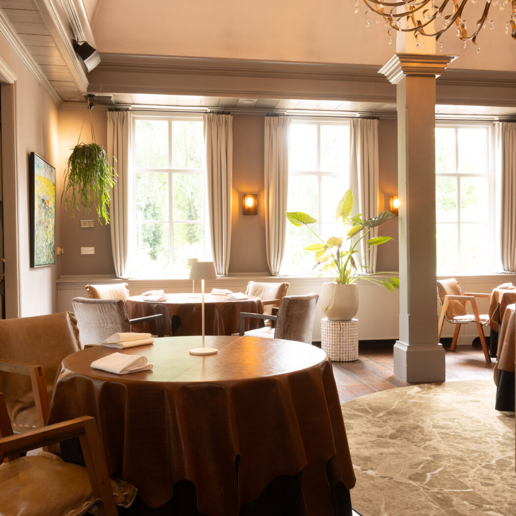 Elegant restaurant interieur met ronde tafels, bruine kleden, beige stoelen, grote ramen en warm licht.