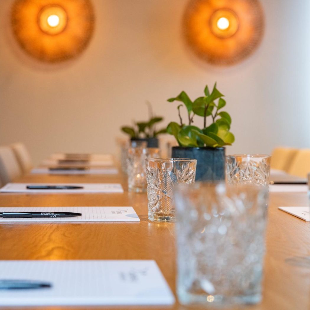Een lange houten vergadertafel, gedekt met notitieblokken, pennen, glazen en groene planten, verlicht door twee wandlampen.