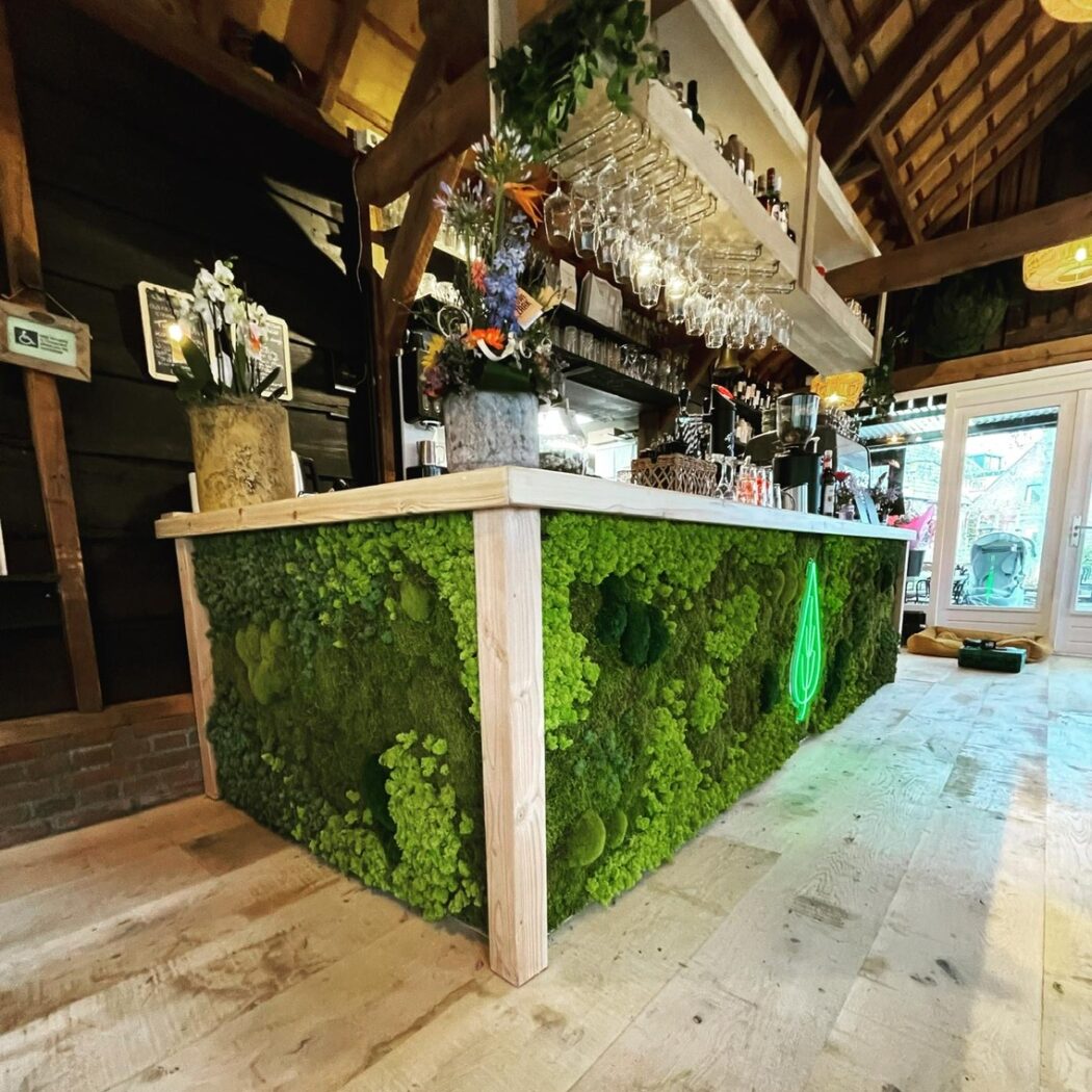 Opvallende bar met groene moswand en een lichtgevend bladlogo, in een sfeervolle ruimte met houten balken en vloer.