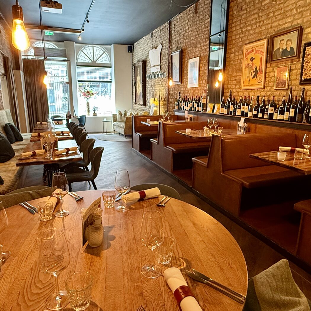 Restaurantinterieur: houten tafels, lederen banken, wijnrek en bakstenen muur.