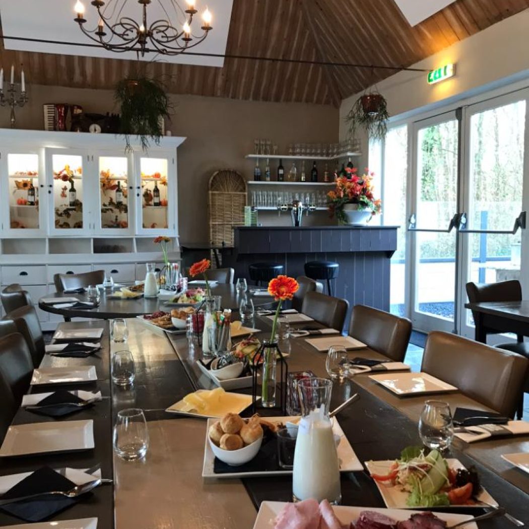 Lange houten tafel rijk gedekt met ontbijtgerechten, kaas, vleeswaren en oranje gerberas in een lichte eetzaal.