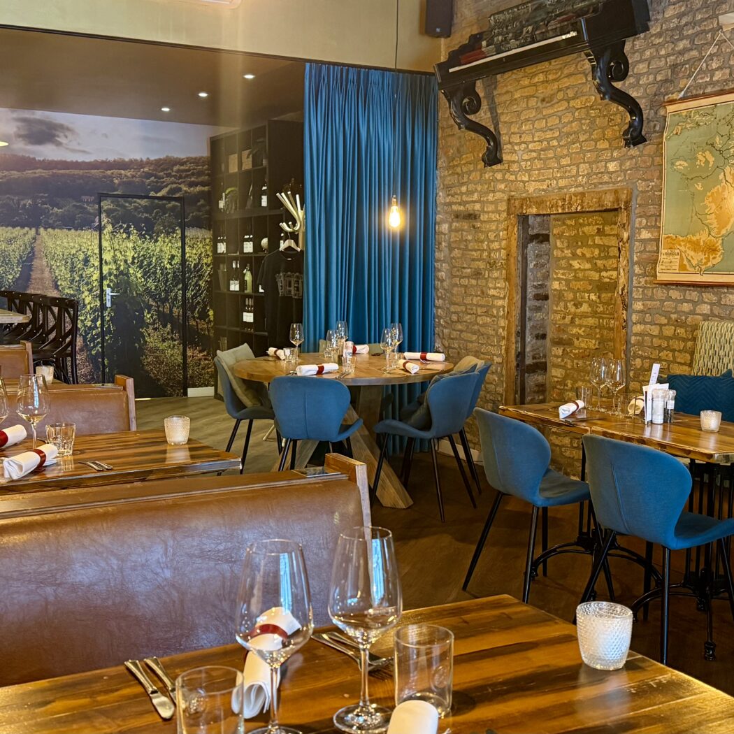 Restaurant interieur: houten tafels, blauwe stoelen, wijnrek en wijngaardmuurschildering.