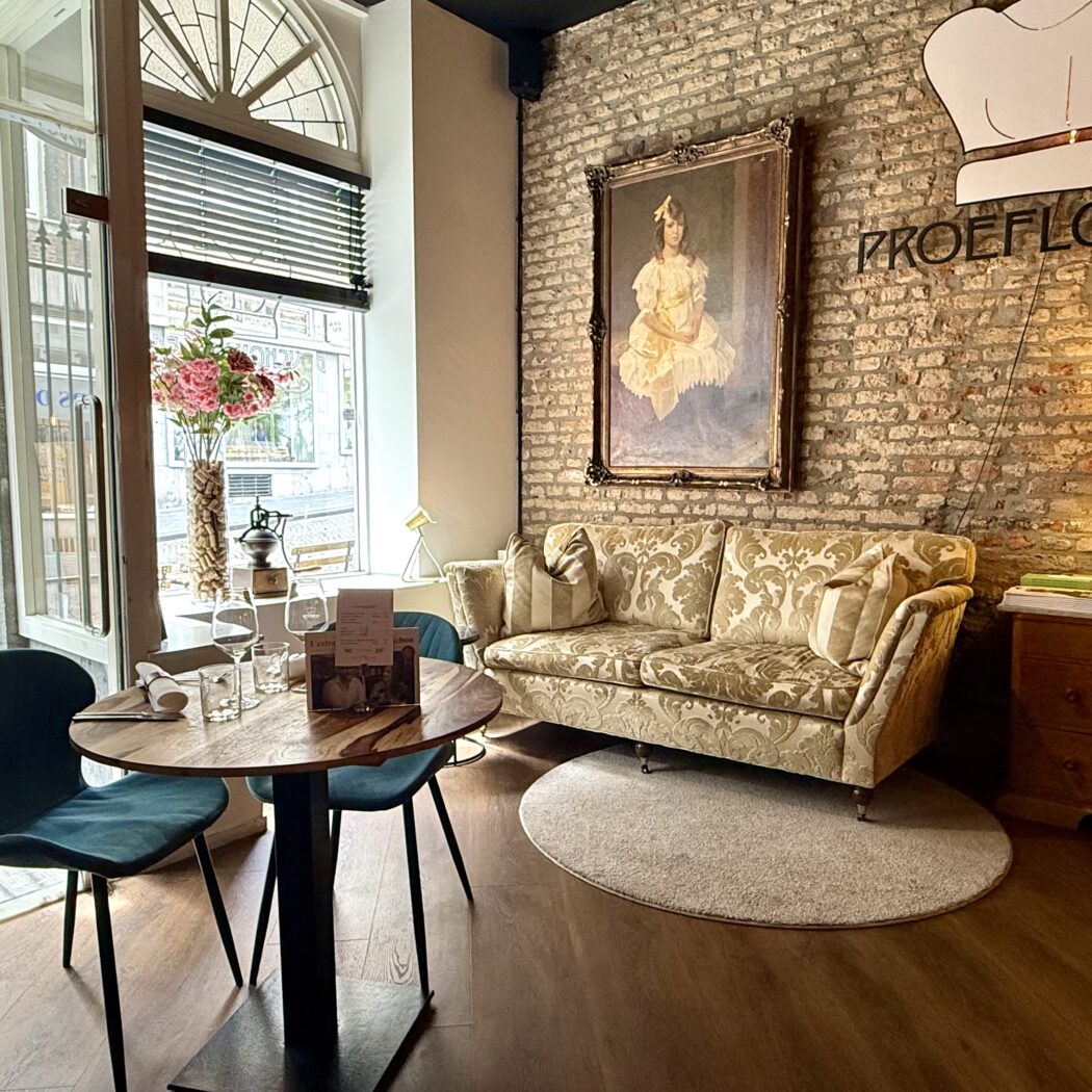 Restaurantinterieur: tafeltje met twee stoelen, gouden sofa, schilderij, bakstenen muur, en Proeflokaal logo.