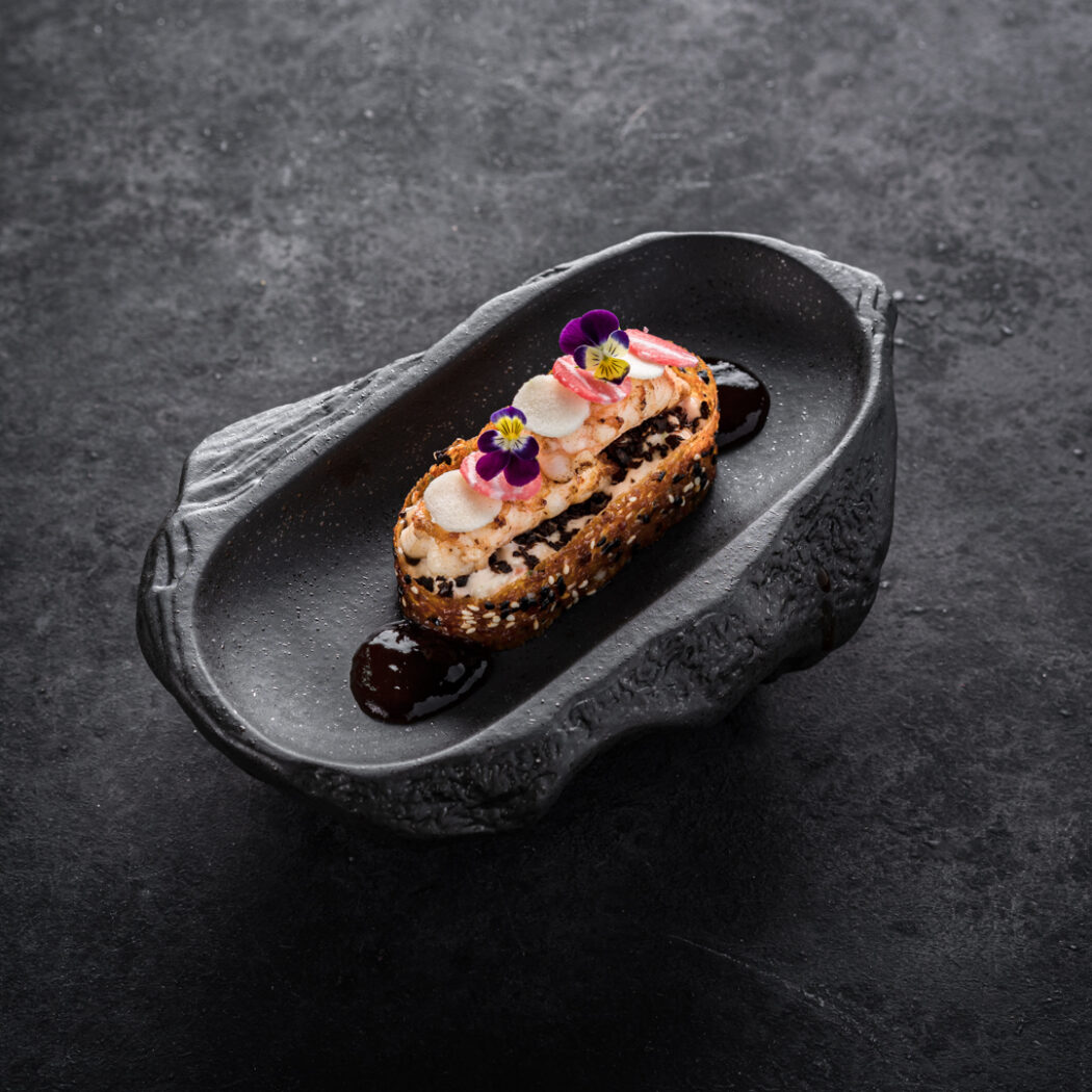 Langwerpig dessert met witte crème, donkere kruimels, roze garnituur en paarse bloemetjes op een uniek donker bord.