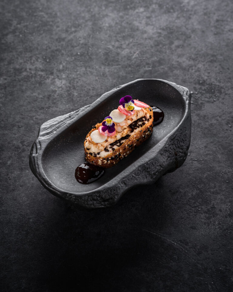 Langwerpig dessert met witte crème, donkere kruimels, roze garnituur en paarse bloemetjes op een uniek donker bord.