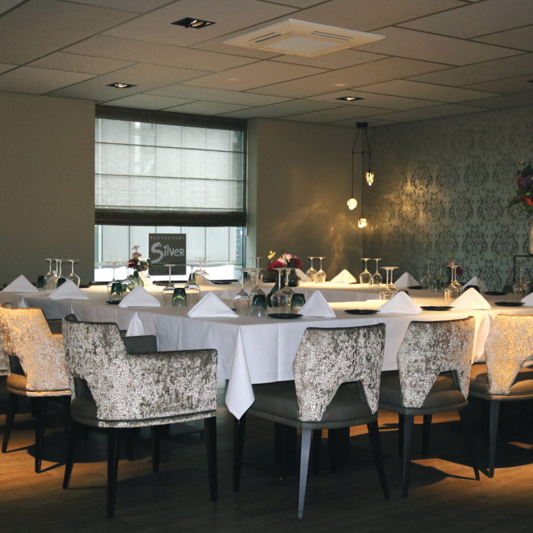 Elegant gedekte lange tafel met glazen, servetten en bloemen in restaurant Silver, omringd door stoelen met patroonbekleding.