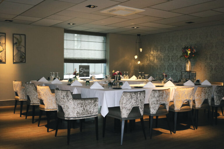 Elegant gedekte lange tafel met glazen, servetten en bloemen in restaurant Silver, omringd door stoelen met patroonbekleding.