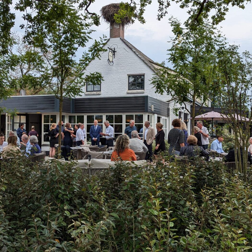 Gasten socialiseren op een buitenterras bij een wit pand met donkere uitbouw en een ooievaarsnest op de schoorsteen.