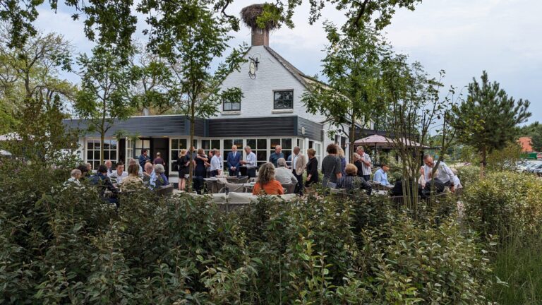Gasten socialiseren op een buitenterras bij een wit pand met donkere uitbouw en een ooievaarsnest op de schoorsteen.