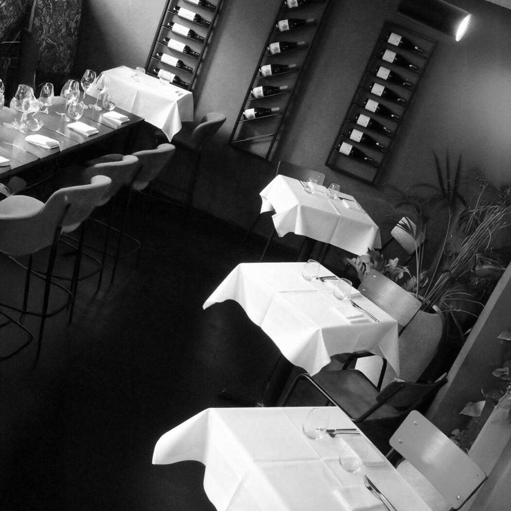 Zwart-wit foto van een restaurant met gedekte tafels, een lange eettafel en wijnrekken vol flessen.