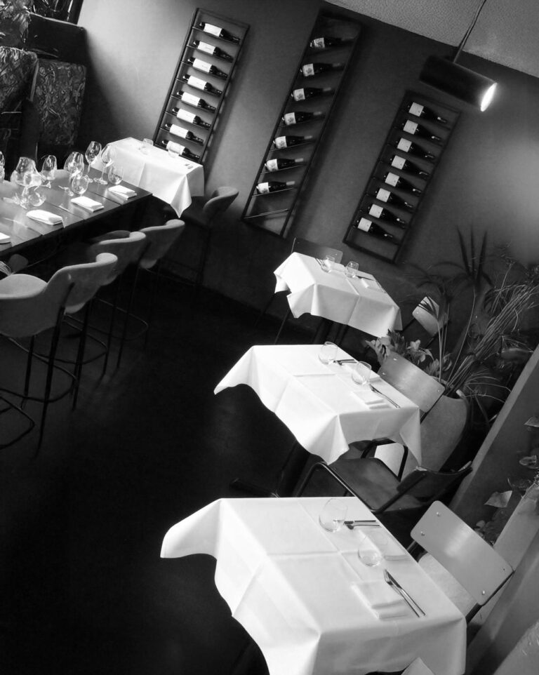 Zwart-wit foto van een restaurant met gedekte tafels, een lange eettafel en wijnrekken vol flessen.