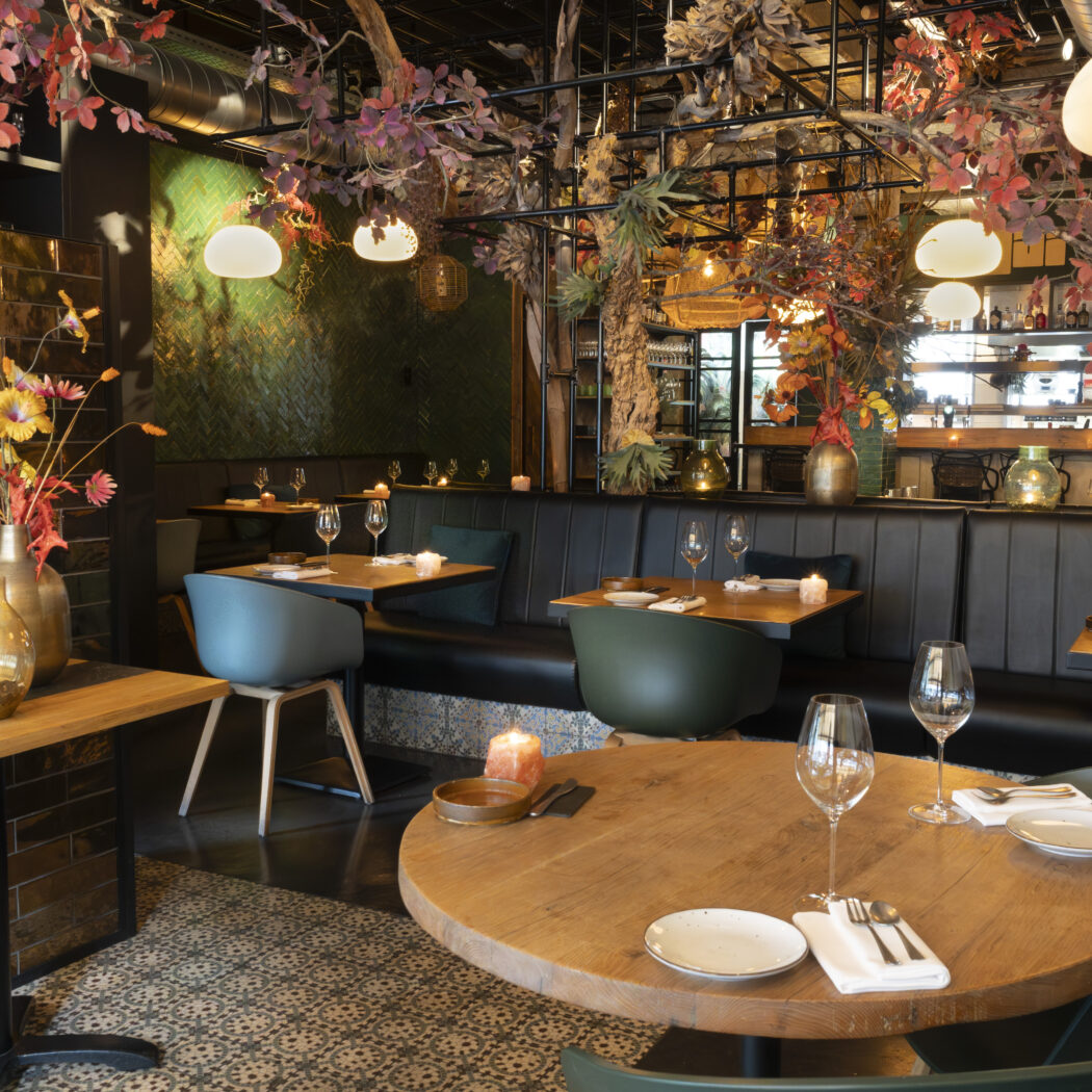 Sfeervol restaurantinterieur met houten tafels, gestoffeerde banken en groene/blauwe stoelen onder herfstblad decoratie.