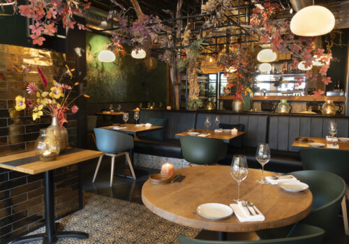 Sfeervol restaurantinterieur met houten tafels, gestoffeerde banken en groene/blauwe stoelen onder herfstblad decoratie.