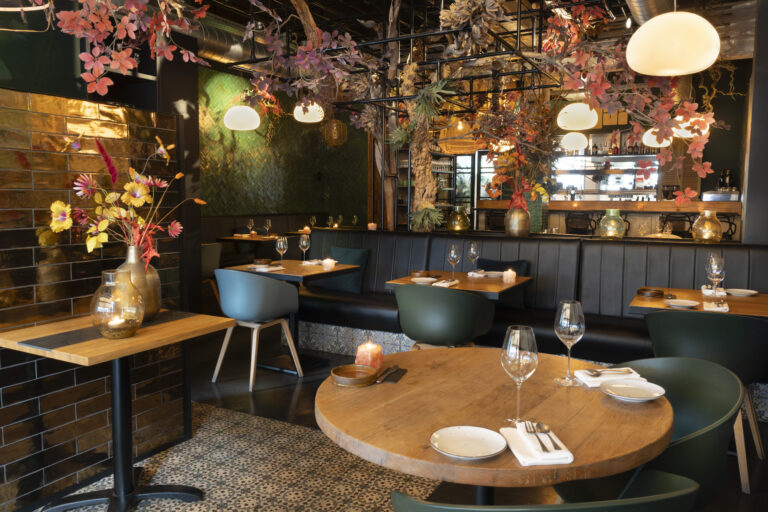 Sfeervol restaurantinterieur met houten tafels, gestoffeerde banken en groene/blauwe stoelen onder herfstblad decoratie.
