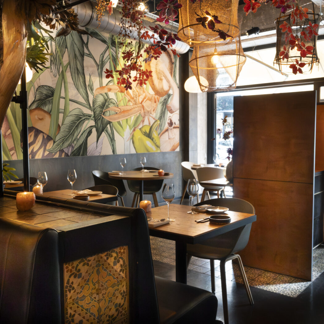 Warm restaurantinterieur met tropische muurschildering, een lange bank, houten tafels en gevlochten hanglampen.