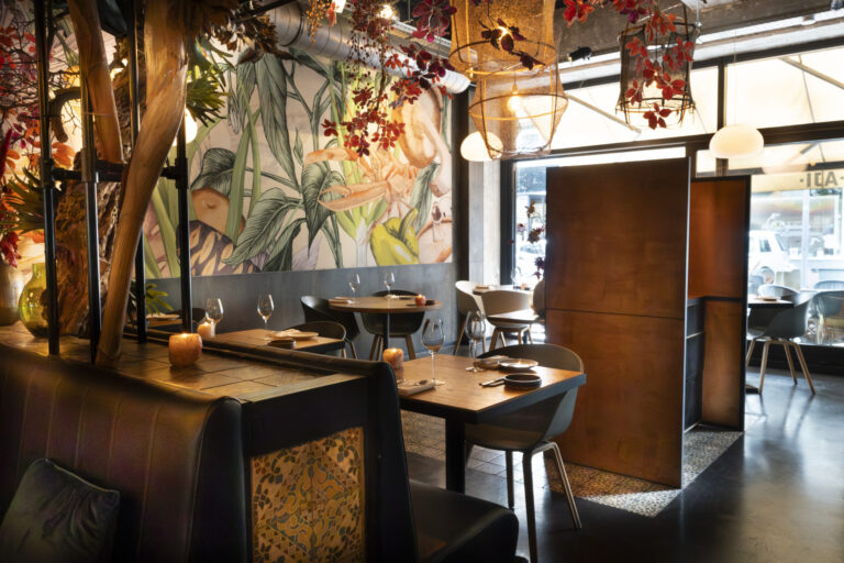 Warm restaurantinterieur met tropische muurschildering, een lange bank, houten tafels en gevlochten hanglampen.