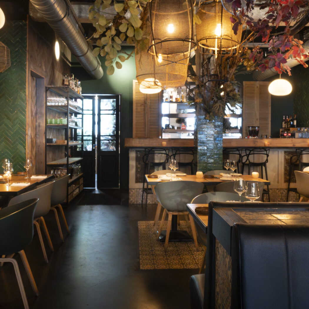 Sfeervol restaurant interieur met houten tafels, groene visgraat tegels, en een bar met hangende planten.