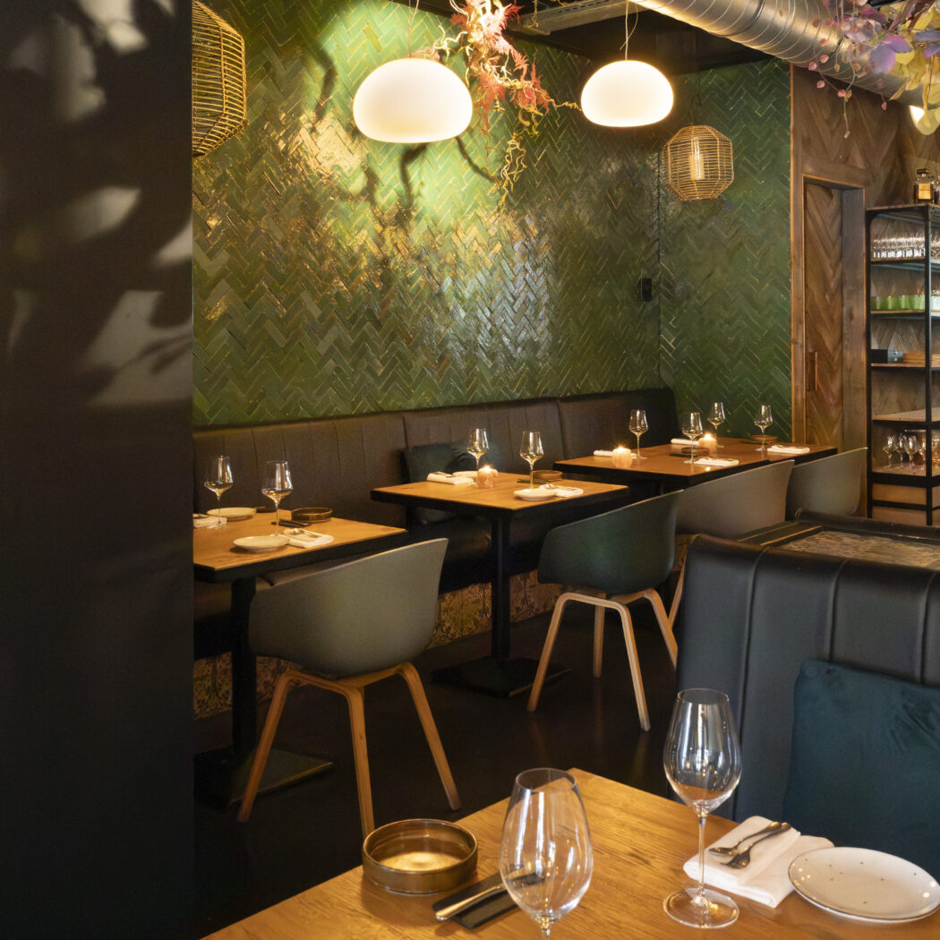 Modern restaurantinterieur met groene visgraattegelwand, gedekte tafels en wijnkoeler.