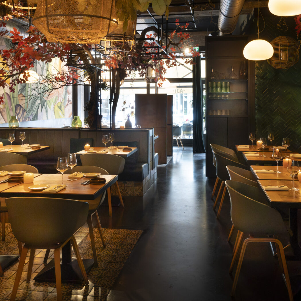 Warm verlicht restaurant met gedekte houten tafels, omringd door weelderige planten, rode bladeren en een botanische muurschildering.