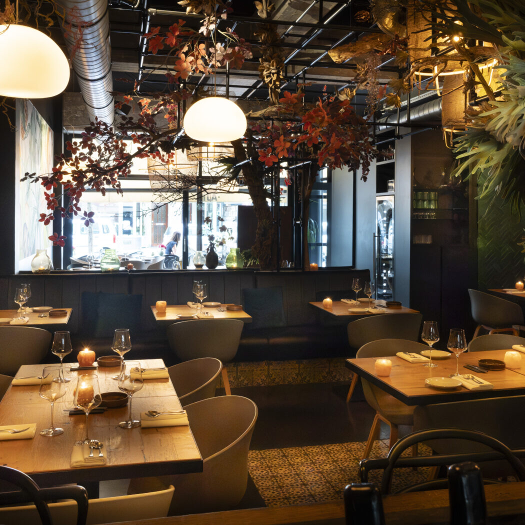 Warm verlicht restaurant interieur met gedekte houten tafels, kaarsen en decoratieve herfstbladeren aan het plafond.