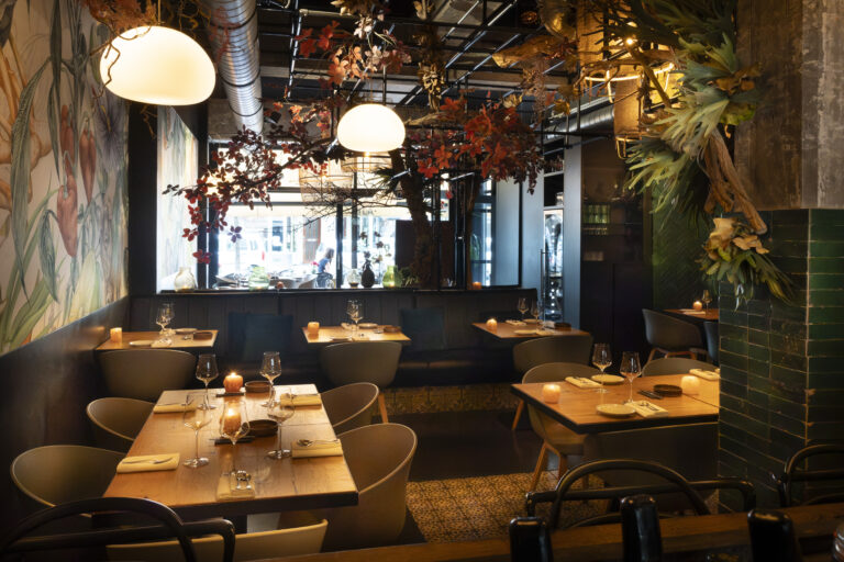 Warm verlicht restaurant interieur met gedekte houten tafels, kaarsen en decoratieve herfstbladeren aan het plafond.