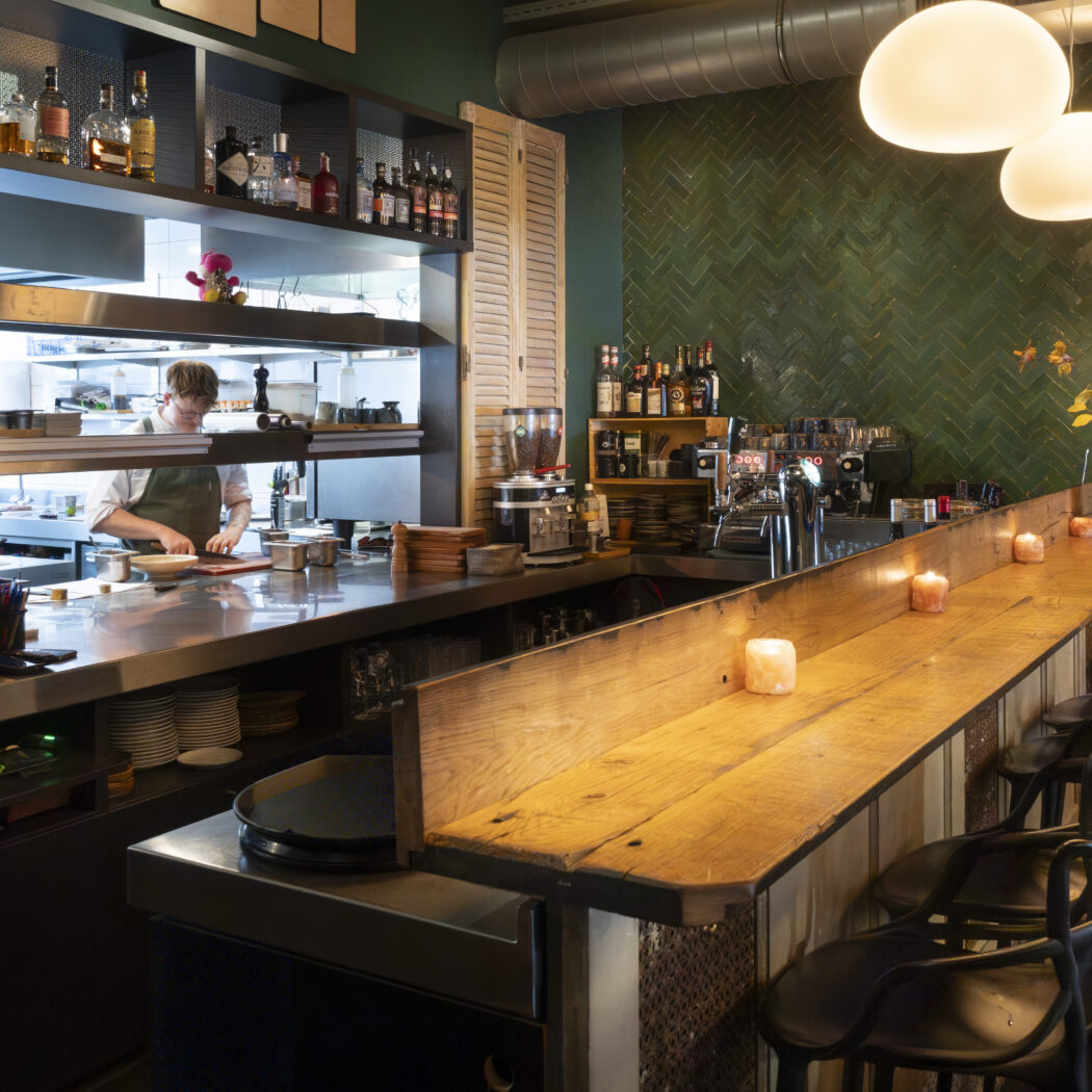 Chef in open keuken van modern restaurant met houten bar, barkrukken, brandende kaarsen en groene tegelwand.