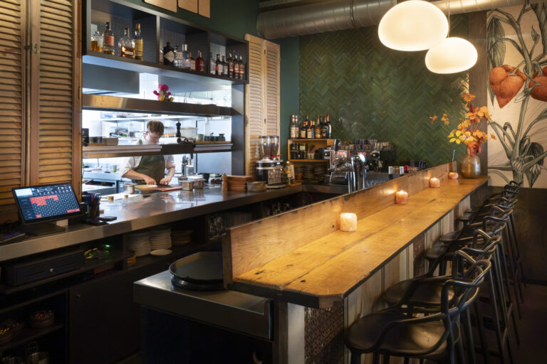 Chef in open keuken van modern restaurant met houten bar, barkrukken, brandende kaarsen en groene tegelwand.