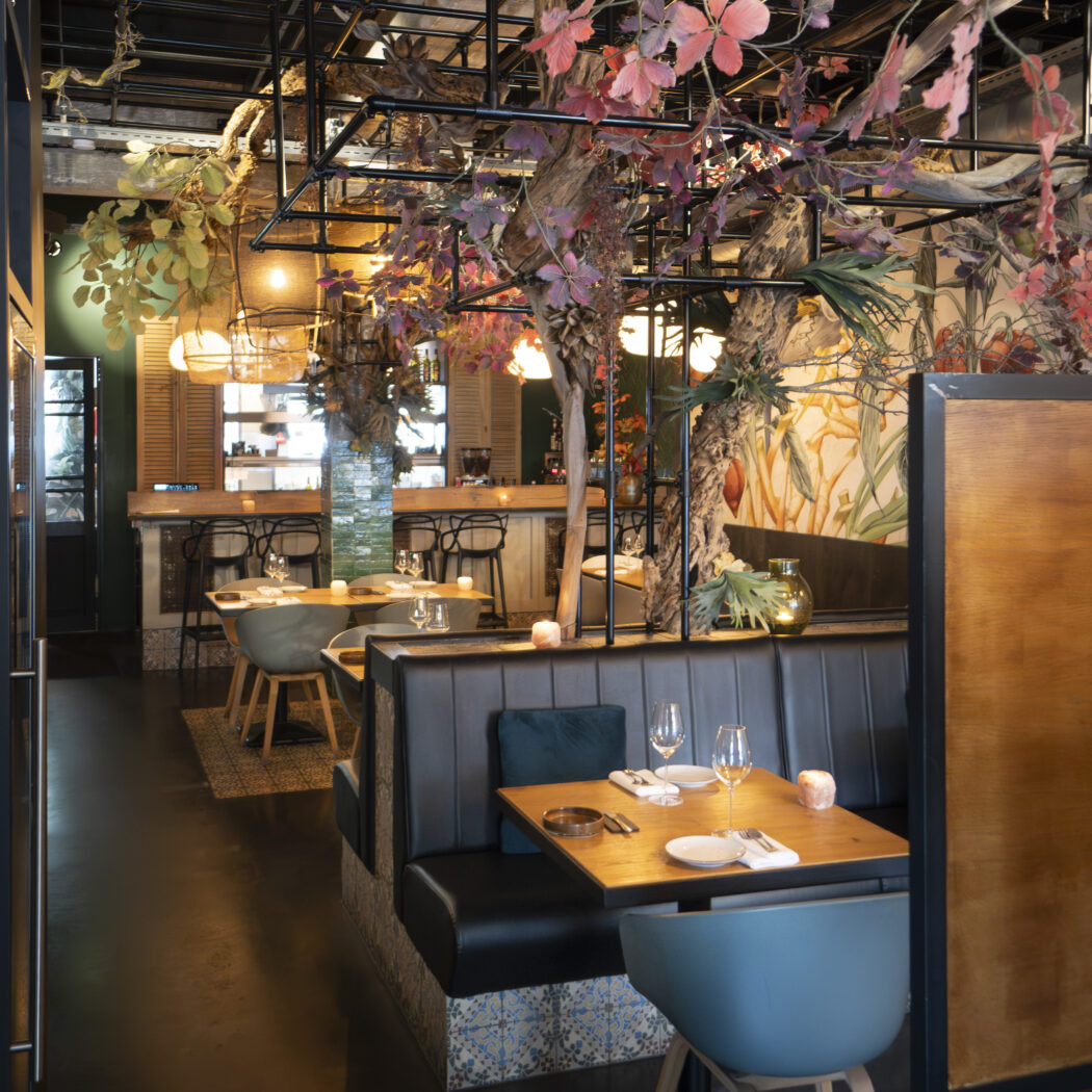 Sfeervol restaurantinterieur met weelderige rode bladeren, leren banken en houten tafels.