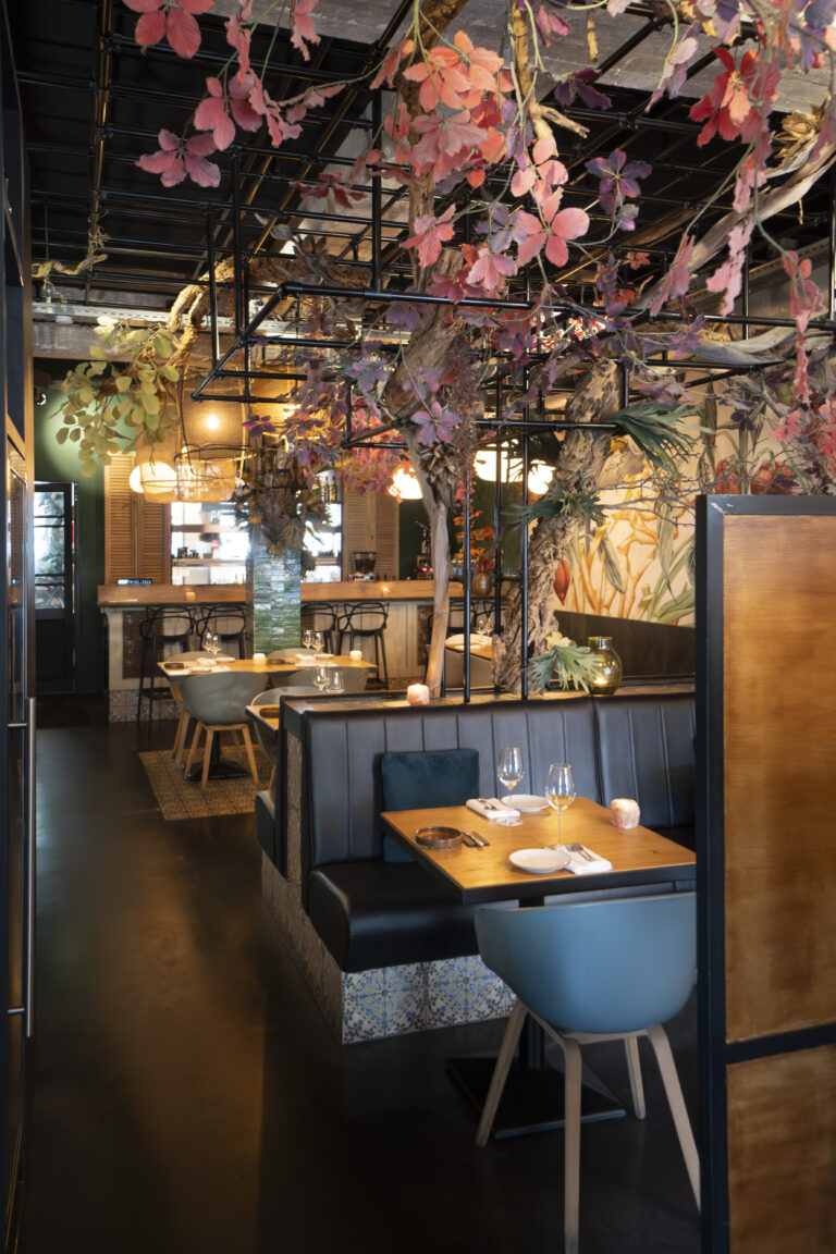 Sfeervol restaurantinterieur met weelderige rode bladeren, leren banken en houten tafels.