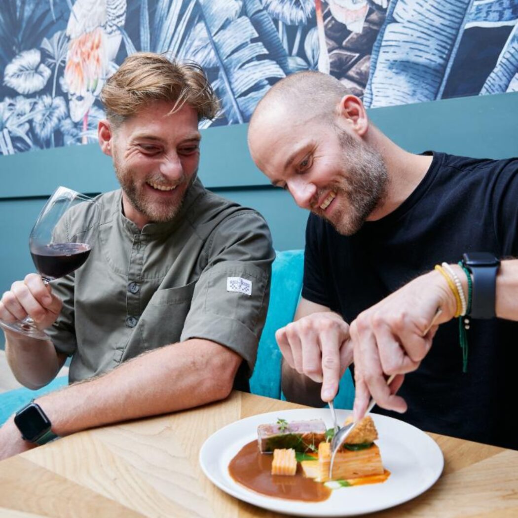 Een lachende chef-kok drinkt rode wijn terwijl een man een gastronomisch gerecht proeft in een restaurant.
