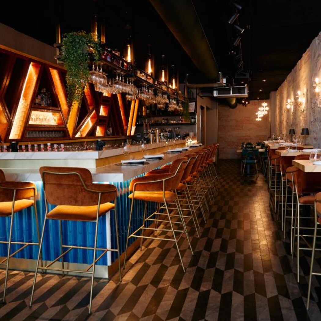 Sfeervol modern restaurantinterieur met een lange bar, oranje fluwelen barkrukken en een verlicht houten wanddecor.